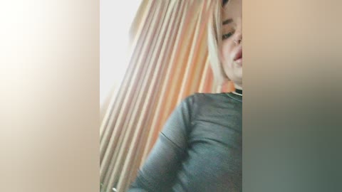 Video of a blonde woman with fair skin, wearing a gray long-sleeved top, partially obscured by a beige curtain.