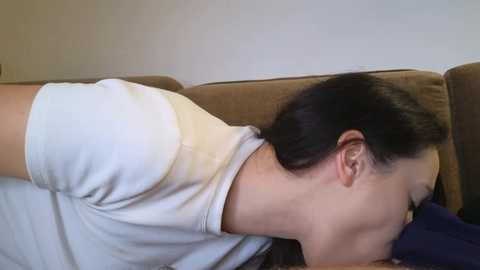 Video of a woman with long black hair, lying on a brown couch with a white shirt, eyes closed, head resting on a dark blue pillow.