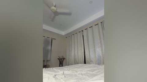 Video of a modern, minimalist bedroom with beige walls, white bed, and white curtains. Ceiling fan, bedside table, and mirror on the left.