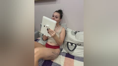A video of a woman with fair skin, dark hair in a bun, wearing a beige tank top and red shorts, sitting on a bed with a patterned blanket, taking a selfie with a tablet.
