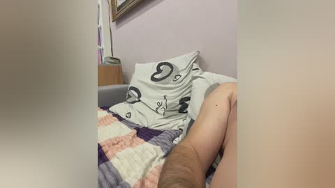 Video of a man lying on a bed with a white and pastel-patterned quilt, his legs visible and muscular, in a softly lit, pastel-colored room with a wooden nightstand and framed artwork on the wall.