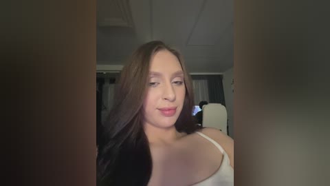 Video of a young Caucasian woman with long, straight brown hair and fair skin, wearing a white spaghetti-strap tank top, smiling subtly in a dimly lit, modern room with dark curtains and a white ceiling fan.