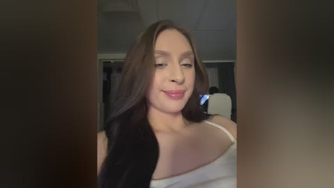 Video of a young woman with light skin and long brown hair, wearing a white off-shoulder top, smiling, standing indoors with blurred background.