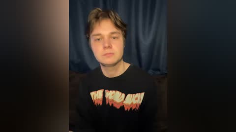 Video of a young man with light skin and brown hair, wearing a black Metallica shirt with flames and red letters, against dark blue curtains.
