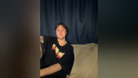 Video of a young man with short hair, wearing a black t-shirt with a graphic design, sitting on a beige couch against a dark blue curtain background.