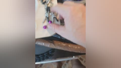 A close-up video of a hand sewing a beige leather shoe, with purple beads attached to the shoe's tongue. The background is blurred, showing a neutral-toned surface.