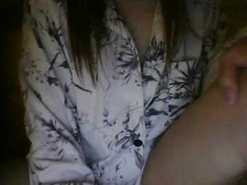 A video of a person's torso, wearing a white floral-patterned shirt. The background is dark and indistinct. The person's hand is visible, holding a beverage. The image is dimly lit, creating a soft, intimate atmosphere.