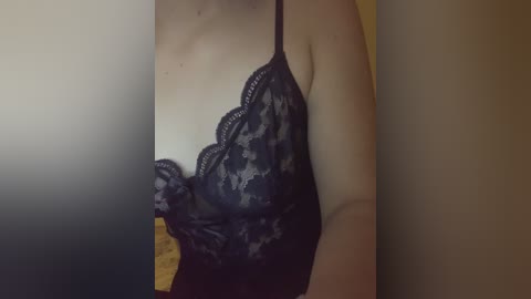 Video of a fair-skinned woman wearing a black lace camisole with scalloped edges, partially visible in dim lighting. Her upper body and arms are prominently displayed, with a blurred background suggesting an indoor setting.