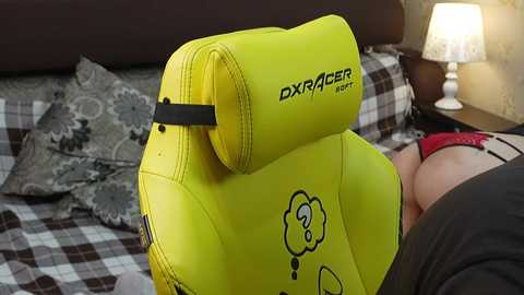 Video of a bright yellow Dxracer gaming chair with a black band and cloud logo, positioned on a bed with a checkered blanket, featuring a partially visible person's leg in red socks.