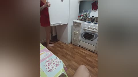 Video of a small kitchen with a woman in a red apron and a man's bare legs, both looking at a white stove.