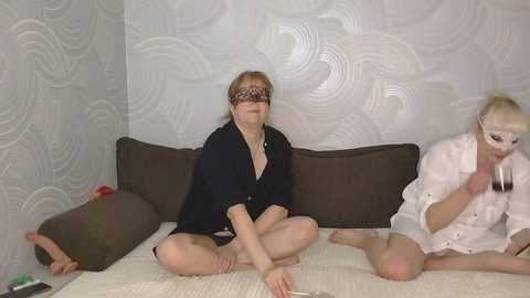 Video of three women in a bedroom with textured wallpaper. One is blindfolded, wearing a black robe, sitting cross-legged on a bed. Another woman, wearing a white robe, is on the right, holding a piece of paper.
