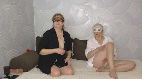 Video of two women, one blindfolded and wearing a robe, the other in a white mask and robe, sitting on a bed with white and brown pillows, patterned wallpaper in the background.