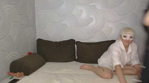 Video of a white woman with platinum blonde hair, wearing a white shirt and a white mask, kneeling on a white bed with brown pillows in a room with white, wavy wallpaper.