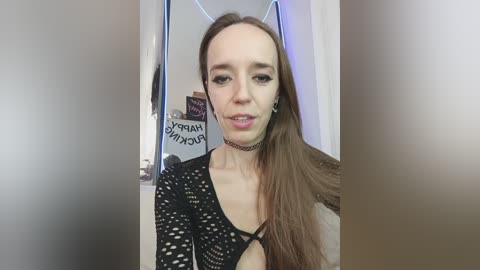 Video of a young Caucasian woman with long brown hair, wearing a black crochet top and choker, standing in a dimly lit room with a mirror and wall decor in the background.
