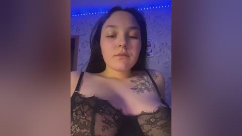 Video of a fair-skinned woman with long black hair, wearing a black lace bra, in a dimly lit room with blue LED strip lights.
