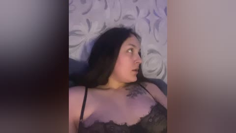 Video of a young woman with long black hair, wearing a black lace bra, lying on a bed with floral-patterned sheets.