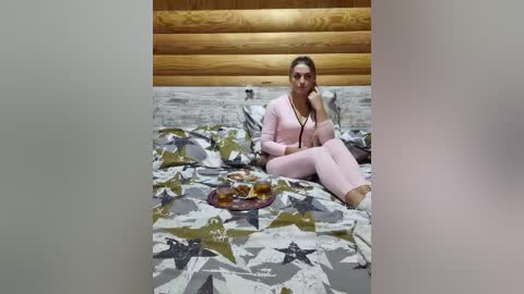 Video of a blonde woman in a pink tracksuit, seated on a bed with a geometric-patterned duvet. She's on a wooden-paneled wall, holding a plate of cookies, in a cozy, rustic room.