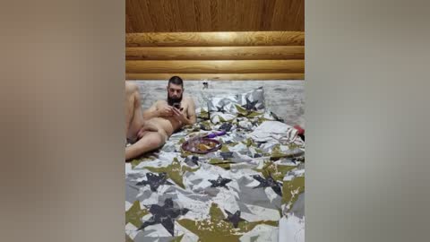 Video of a bearded, nude man with a medium build, lying on a bed with camouflage-patterned bedding, inside a rustic cabin with wooden walls and ceiling.