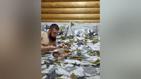 Video of a bearded, shirtless man with a muscular build, focused on a tray of food and a drink on a bed with a patterned duvet in a rustic wooden room.