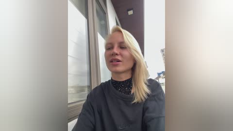 Video of a blonde woman in a black robe, sitting in a dimly lit hallway with glass windows. She appears contemplative, with a soft-focus background showing an indistinct cityscape.