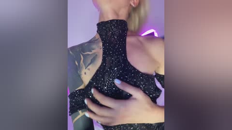 Video of a woman with short blonde hair, wearing a black, glittery, off-the-shoulder top, showcasing a detailed tattoo on her arm. She is holding her breasts with manicured nails. Background is blurred, with purple lighting.