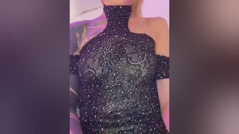 Video of a woman wearing a black, sparkly, off-the-shoulder dress with intricate embroidery, set against a blurred, colorful background. Her fair skin and blonde hair are partially visible.