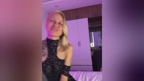 Video of a smiling, light-skinned blonde woman in a black sequined halter top, sitting on a bed in a modern bedroom with purple lighting.