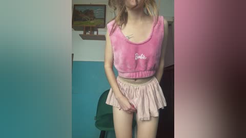 Video of a slender, fair-skinned woman with shoulder-length blonde hair, wearing a pink cropped top and a short, ruffled pink skirt. She stands in a room with teal and white walls, a green chair, and a framed painting.