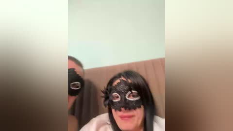 A video captures a man with a black mask, standing behind a woman with long black hair and a similar mask. The background features a pale green wall and a wooden panel.