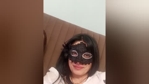 Video of a young woman with long black hair, wearing a black lace masquerade mask with white eyes, and a white top, seated on a wooden bench against a light blue wall.