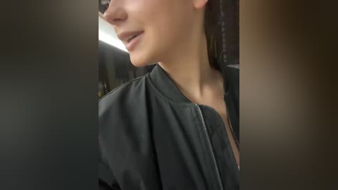 Video of a woman's profile with fair skin, wearing a black zip-up jacket, smiling subtly, in a dimly lit room with blurred background.