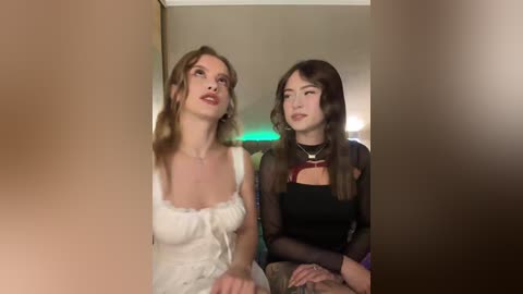 Video of two young women with long hair, one in a white dress, the other in a black top, seated indoors with a blurred background.