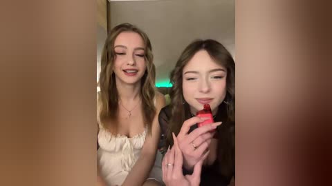 Video of two young women with fair skin and long hair, one wearing a white dress, the other a black top, eating red gummy candy. Background shows a blurred room with green lighting.