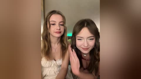 Video of two young women, one with long, wavy hair in a cream dress, the other with long, straight hair in a pink dress, both smiling and looking at a phone.