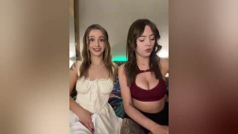 Video of two young women sitting on a bed, one in a white dress, the other in a burgundy bralette, both with fair skin, smiling, indoors with blurred background.