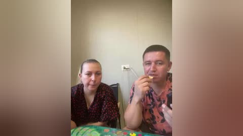 Video of a middle-aged man and woman in a dimly-lit room, both eating a sandwich, with a blurred background. The man has short hair, wears a patterned shirt, and holds a fork. The woman has blonde hair tied back, wears a dark blouse, and holds a knife.
