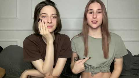 Video of two young women with long hair, one with dark hair, one with light brown hair, both wearing casual shirts, sitting side by side on a couch.