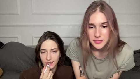 Video of two young women, one with long brown hair and another with long light brown hair, sitting on a gray couch, using a lipstick. Background features a white-paneled wall.
