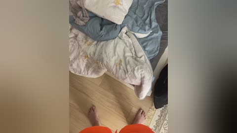 Video of a person standing barefoot on light wood floor, wearing orange shorts, looking down at a messy bed with a white and blue blanket and a white pillow.