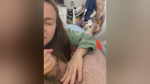 Video: A young woman with long brown hair, wearing a green sweater, leans over a man lying on a bed. He's wearing red shorts. The room is messy with clothes and bags.