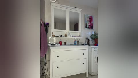A video of a modern, well-lit bathroom featuring a white vanity with two drawers, a mirror, and a purple towel hanging on a wall.
