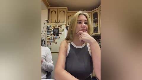 Video of a young blonde woman with medium build, wearing a grey sleeveless top, holding her chin in a kitchen with light-colored wooden cabinets, a refrigerator covered in magnets, and another person partially visible.