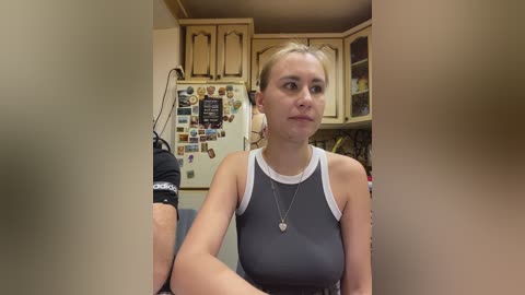 Video of a blonde woman in a grey tank top, seated in a kitchen with wooden cabinets, fridge adorned with photos, and a black bra hanging on the left.