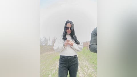 Video of a woman with long black hair, wearing sunglasses and a white sweater, standing in a foggy, grassy field with another person partially visible.