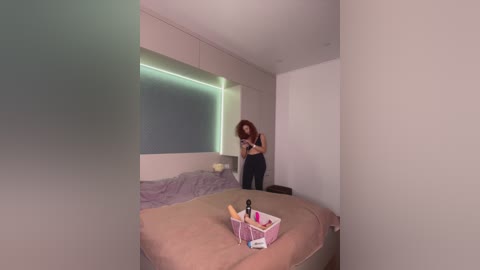 Video of a minimalist bedroom with a woman in black, taking a selfie in front of a beige bed with a pink storage box on it.