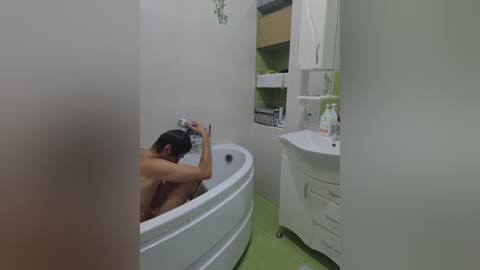 A video of a naked man with dark hair, seated in a white bathtub, using a smartphone. The background shows a white sink and green-tiled wall in a modern bathroom.