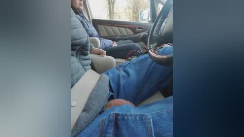 Video of a person in a car, wearing a gray sweater and blue jeans, seated in the passenger seat with a child in a car seat on their lap.
