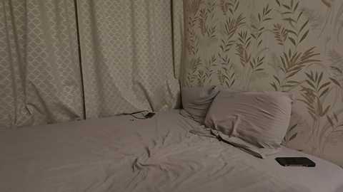 Video of a messy bedroom with rumpled bed sheets, a pillow, and a phone on the bed, set against a wall with floral wallpaper.