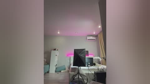 Video of a modern, dimly-lit bedroom with a grey carpet, a desk with a monitor and keyboard, a white dresser, and a large fan.