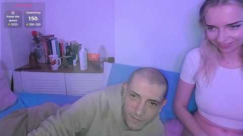 Video of a man with a bald head, light skin, and light brown shirt lying on a blue couch, and a smiling woman with long blonde hair, light skin, and a white top sitting beside him.